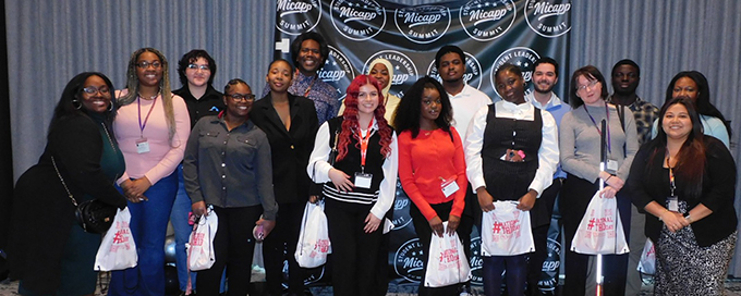 A group of trio students together in front of a Micapp Summit backdrop, smiling for a photograph